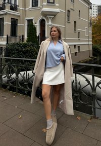 Ανοιχτό μπλε πουλόβερ με V-τελείωμα, λευκή φούστα, μπεζ μακρύ παλτό, γκρι κάλτσες και καφέ πάνινα παπούτσια ενάντια σε φόντο πόλης.