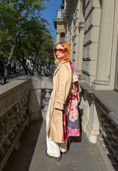 Mujer con pelo rojo que lleva una gabardina beige y gafas de sol rojas, de pie en un balcón de piedra junto a un edificio histórico bajo la luz del sol.