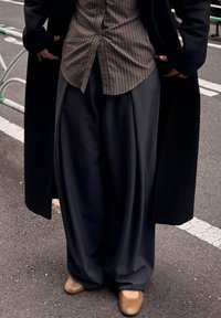 Pantalon large gris foncé avec un devant plissé, associé à une chemise à rayures marron boutonnée et des chaussures à enfiler couleur tan.