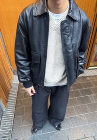 Crna kožna bomber jakna s ovratnikom, dva prednja džepa, preko svijetlo bež pletenog džempera, uparena s širokim crnim hlačama.