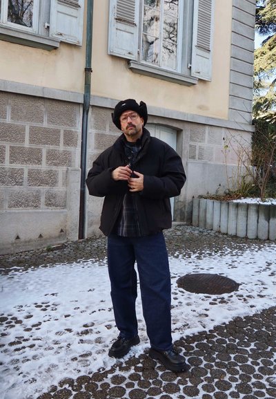 Hombre con chaqueta negra, camisa a cuadros y gorro de piel negro de pie en un camino empedrado nevado frente a un edificio con ventanas con contraventanas.