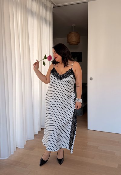 Mujer con vestido negro con lunares blancos oliendo una rosa rosa, de pie junto a cortinas blancas y un suelo de madera en una habitación moderna.