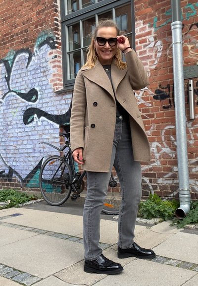 Abrigo de lana beige de doble botonadura con solapas anchas, jeans gris de corte ajustado y zapatos planos negros, frente a una pared de ladrillo con graffiti.