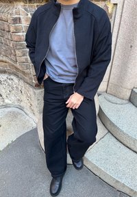 Personne portant un pantalon noir à jambes larges, une veste noire dézippée sur un t-shirt gris, des chaussures noires, debout près d'escaliers en pierre et d'un mur en briques en plein air.