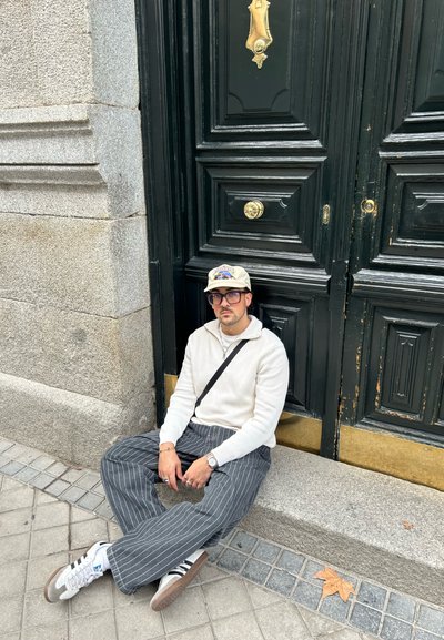 Una persona está sentada en un escalón de piedra al lado de una gran puerta verde oscura con herrajes de latón. Lleva un suéter blanco, pantalones a rayas y zapatillas.