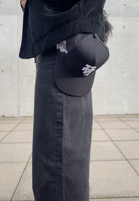 Czarna czapka baseballowa z białym logo, pokazana pod kątem. Noszona z czarną kurtką z korduroy i długą ciemną spódnicą, na tle betonowym.