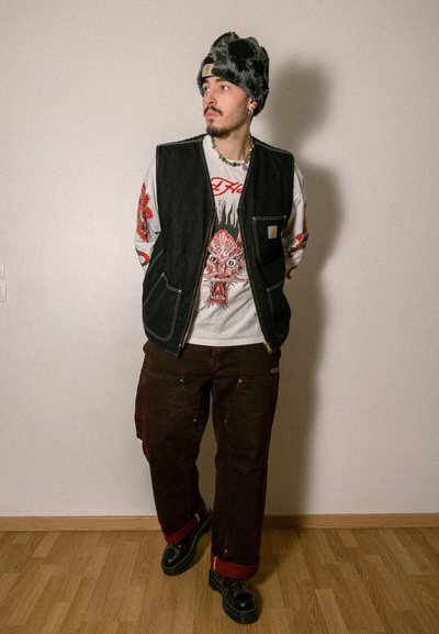 Chaleco de mezclilla negra sobre una camiseta de manga larga blanca con diseño rojo, combinado con pantalones cargo marrones y zapatos negros con costuras amarillas.