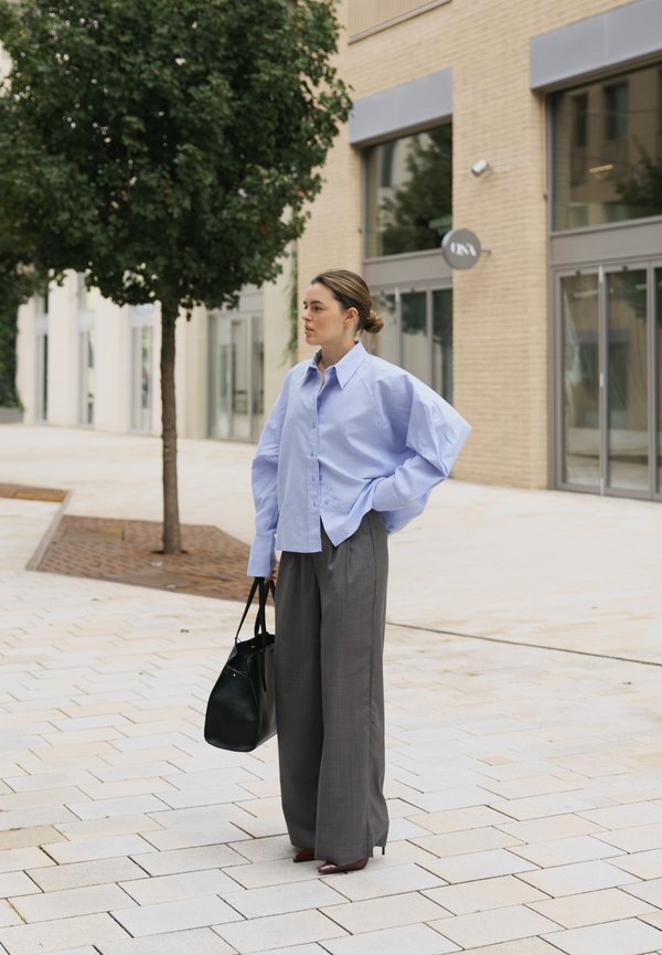 Cămașă oversized de culoare albastru deschis cu mâneci bufante, asortată cu pantaloni gri cu picior larg. Geantă neagră și pantofi maro cu vârf ascuțit. Decor urban.
