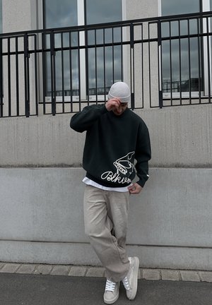 Hombre con gorro gris, sudadera negra con gráfico blanco, pantalones beige y zapatillas blancas, de pie contra una pared de concreto con una barandilla negra.