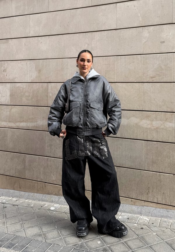 Mlada oseba stoji ob steno iz ploščic, oblečena v prevelik sivi suknjič, široke črne hlače z neti "LOVE" dizajnom in črne platformne čevlje.