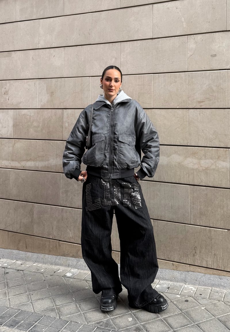 Young person stands against tiled wall wearing oversized gray jacket, loose black pants with studded "LOVE" design, and black platform shoes.