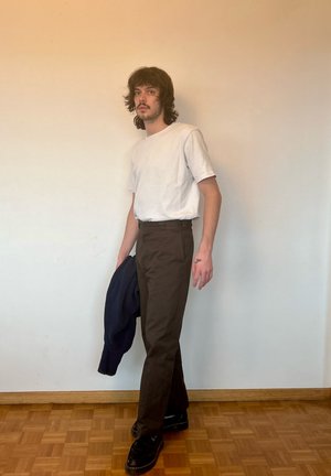 Camiseta blanca de cuello redondo, pantalones ajustados marrones y zapatos negros. Sosteniendo una chaqueta azul marino. Suelo de madera y fondo de pared sencilla.