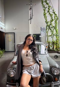 Nainen, jolla on lasit, harmaat shortsit ja bleiseri, nojautuu vintage-harmaaseen autoon modernissa tilassa, jota koristavat kasvit ja roikkuva pyörä.