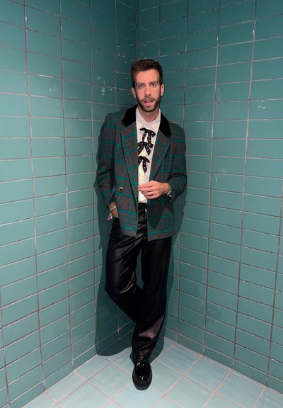 Hombre con blazer de cuadros verdes, camisa blanca con lazos negros, pantalones de cuero negros y zapatos negros apoyado en una esquina con azulejos color verde azulado.