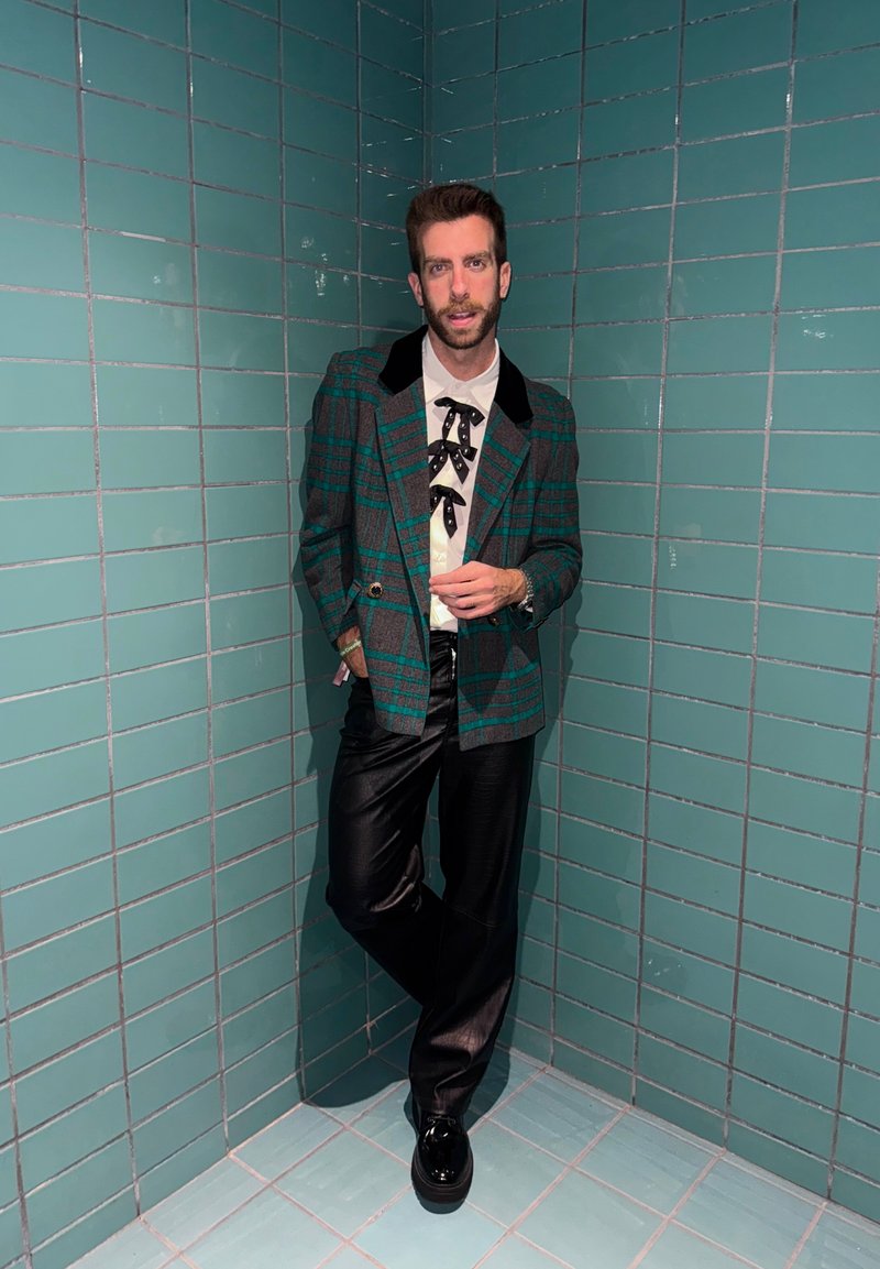 Moški v zeleni karirasti jakni, beli srajci s črnimi metuljčki, črnih usnjenih hlačah in črnih čevljih se opira ob kotno steno obloženo s turkiznimi ploščicami.