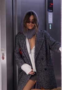 Grau gehäkelter Mantel im Fischgrätenmuster mit übergroßen Revers, über einem weißen Hoodie getragen. Abgerundet wird der Look durch einen grauen Strickschal und eine dunkle Sonnenbrille.