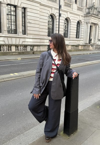 Mujer con blazer gris y camisa a rayas apoyada en un bollardo negro en una calle de la ciudad, con un edificio clásico de piedra al fondo.