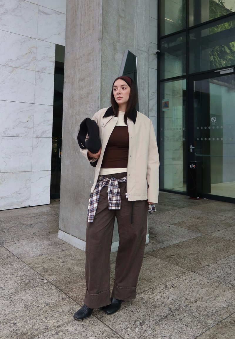 Femme debout près d'un pilier en béton à côté de portes vitrées, portant une veste beige, un pantalon marron, une chemise à carreaux nouée à la taille, tenant un chapeau noir.