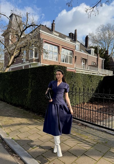 Mujer con vestido azul marino y botas blancas camina por la acera junto a un seto alto y una casa de ladrillo con toldos a rayas rojas bajo un cielo parcialmente nublado.