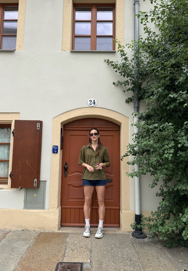 Mujer con gafas de sol, camisa oliva y pantalones cortos, de pie frente a una puerta de madera con el número 24, junto a un árbol frondoso y una ventana con persianas.