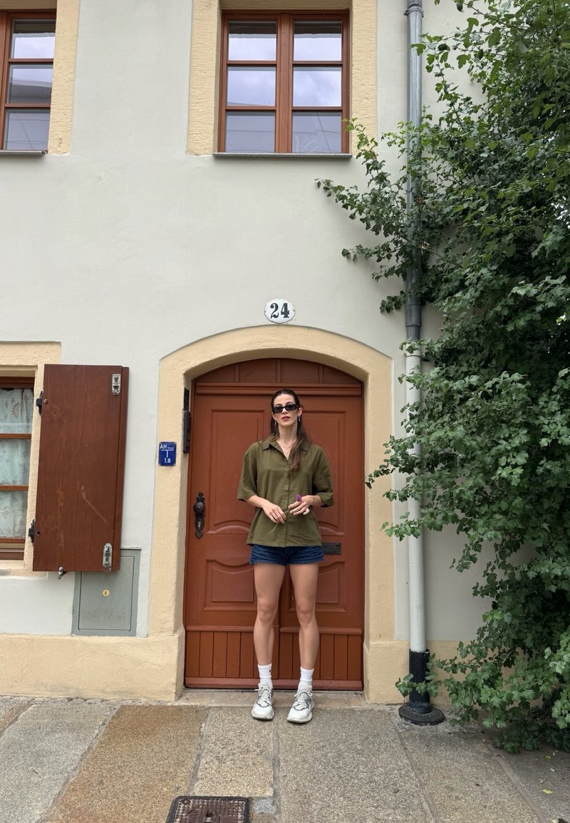 Donna con occhiali da sole, camicia oliva e pantaloncini in piedi davanti a una porta di legno con il numero 24, accanto a un albero frondoso e una finestra con persiane.