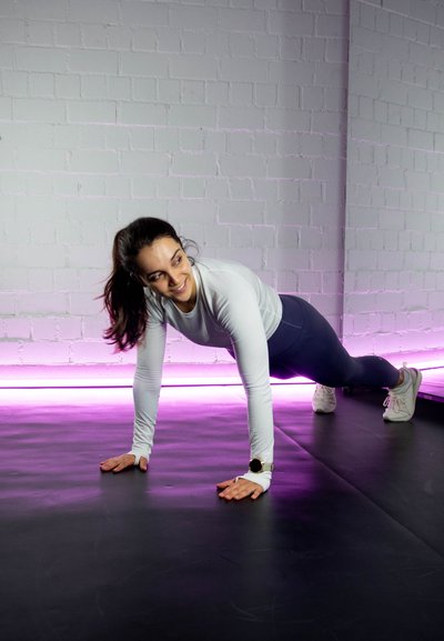 Una mujer realiza una flexión en una esterilla negra, vestida con una camiseta ajustada de manga larga blanca y legging azules, con luces LED rosadas en el fondo.