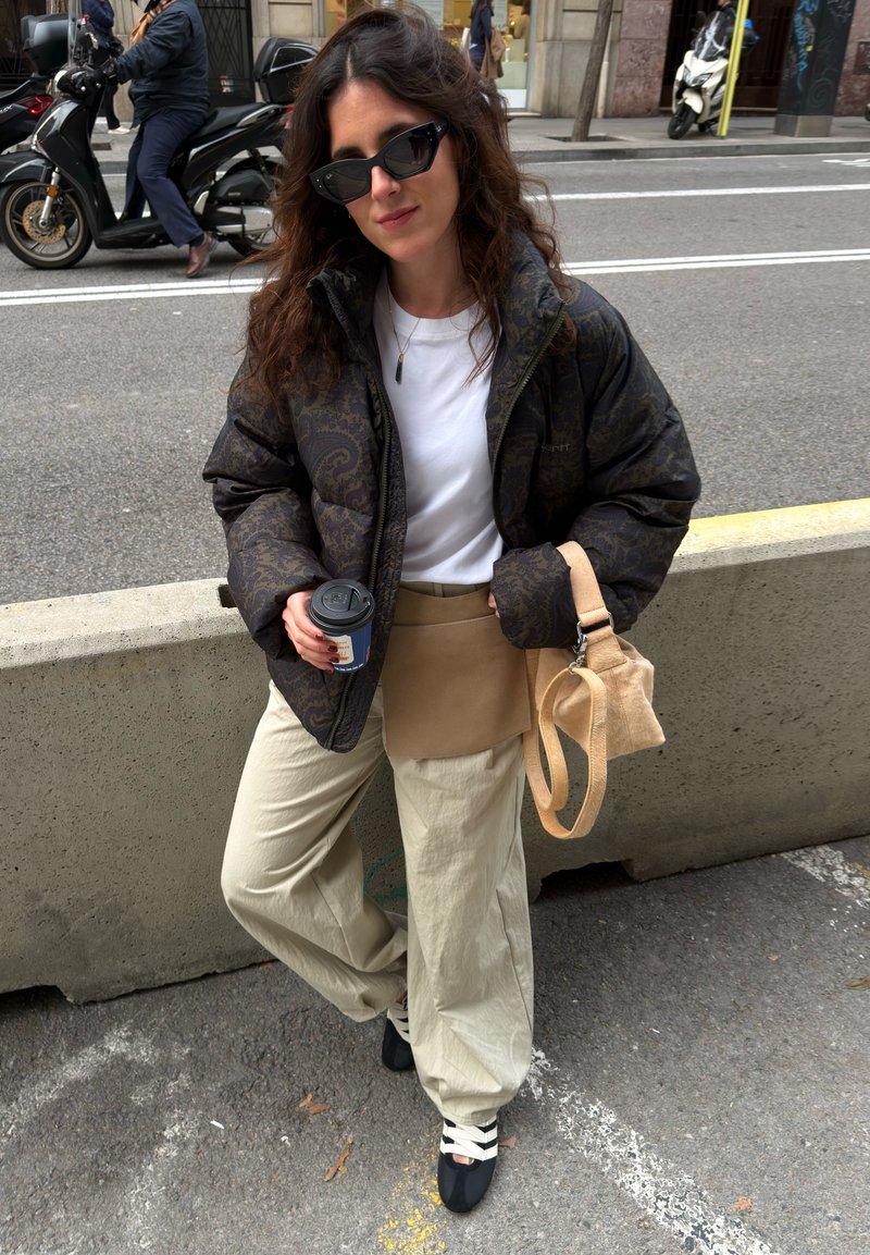 Femme portant une doudoune noir à motif, chemise blanche, pantalon beige, lunettes de soleil noires, tenant une tasse de café à emporter et un sac à main beige dans une rue de la ville.