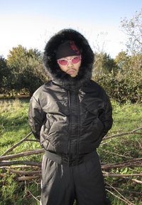 Casaco acolchoado preto com capuz forrado a pelo sintético, usado com um gorro de malha preto e óculos de sol rosa. O fundo apresenta árvores e relva.