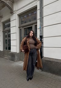 Casaco trench de camurça castanho sobre uma blusa ajustada castanha e jeans largos escuros. Sapatos de salto agudo pretos. Segurando uma bolsa de peluche castanha. Cenário urbano.
