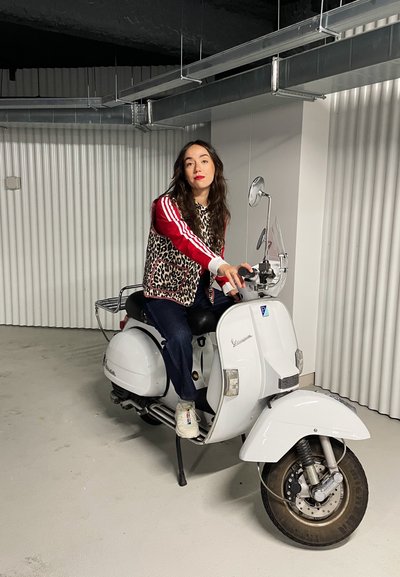 Scooter blanca vintage con acentos de cromo, parte frontal redondeada y un solo espejo lateral. El conductor lleva puesta una chaqueta de estampado de leopardo y jeans.