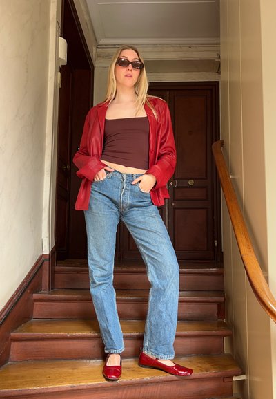 Chaqueta de cuero roja, top corto marrón, jeans de mezclilla azules y zapatos planos rojos. De pie en unas escaleras de madera con una puerta marrón al fondo.