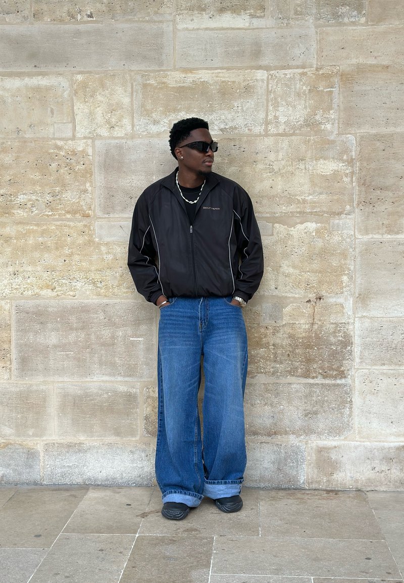 Black zip-up jacket with white accents, black T-shirt, oversized blue jeans, and dark footwear. Set against a light stone wall.