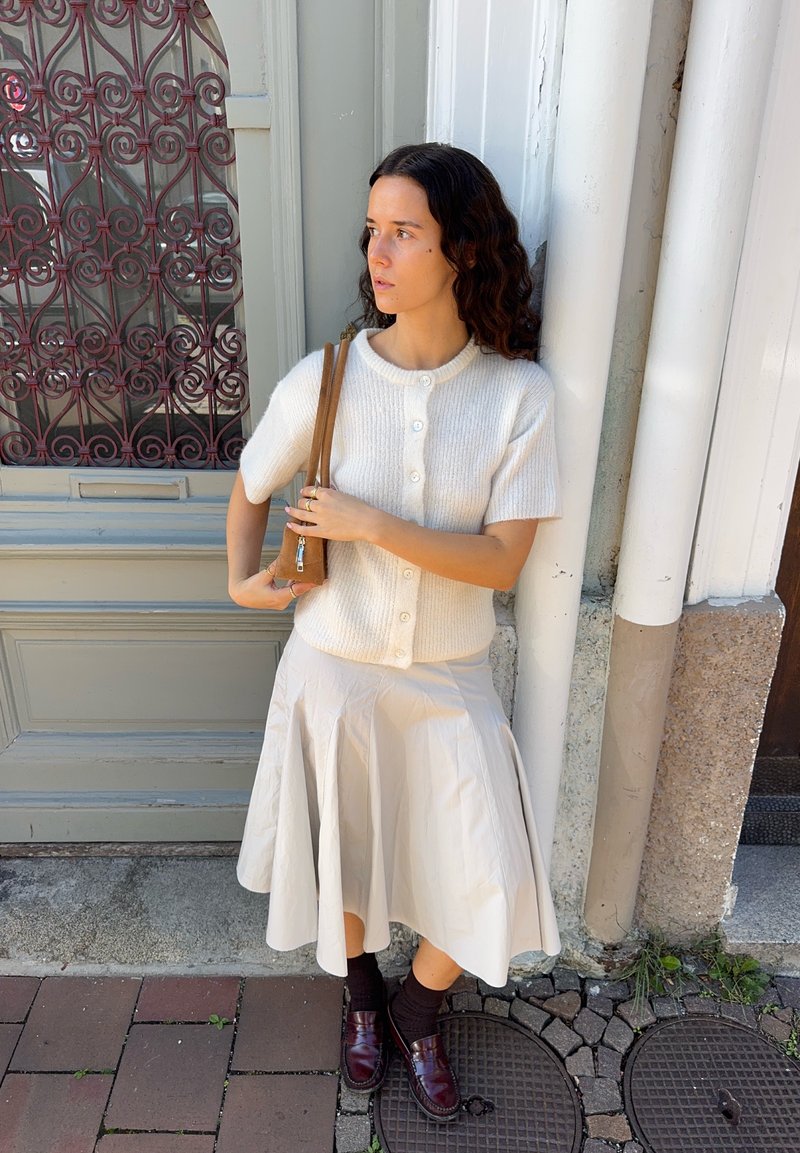 Cardigan corto crema con maniche a sbuffo e bottoni, abbinato a una gonna fluida beige chiaro. Calze marrone scuro e mocassini bordeaux completano l'outfit.
