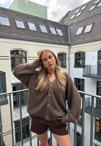 Brązowy kardigan z żeberkami i dużymi guzikami, zestawiony z ciemno-brązowymi szortami w białe paski. Noszony wewnątrz, z widokiem na balkon.