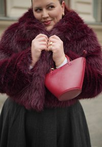 Γυναίκα που φοράει παλτό από μπορντό συνθετική γούνα, κρατώντας το γιακά με τα δύο χέρια, κρατώντας μία τσάντα χειρός σε ταιριαστό μπορντό χρώμα, χαμογελώντας απαλά.