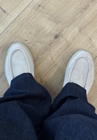 Beige suede loafers with flat soles, featuring a rounded toe and stitch detailing, paired with dark blue denim pants on a wooden floor.