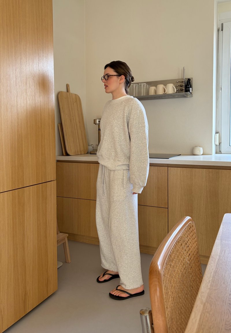 Ženska v sivih trenirkah in jopa, z očali in natikači, stoji v kuhinji z lesenimi omaricami in rezalnimi deskami.