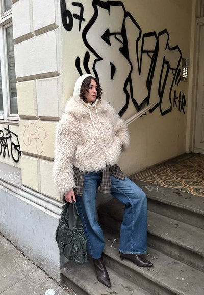 Mujer con una chaqueta con capucha y peluda y vaqueros azules, sosteniendo una bolsa verde oscuro, de pie en unas escaleras al aire libre junto a una pared cubierta de grafitis.