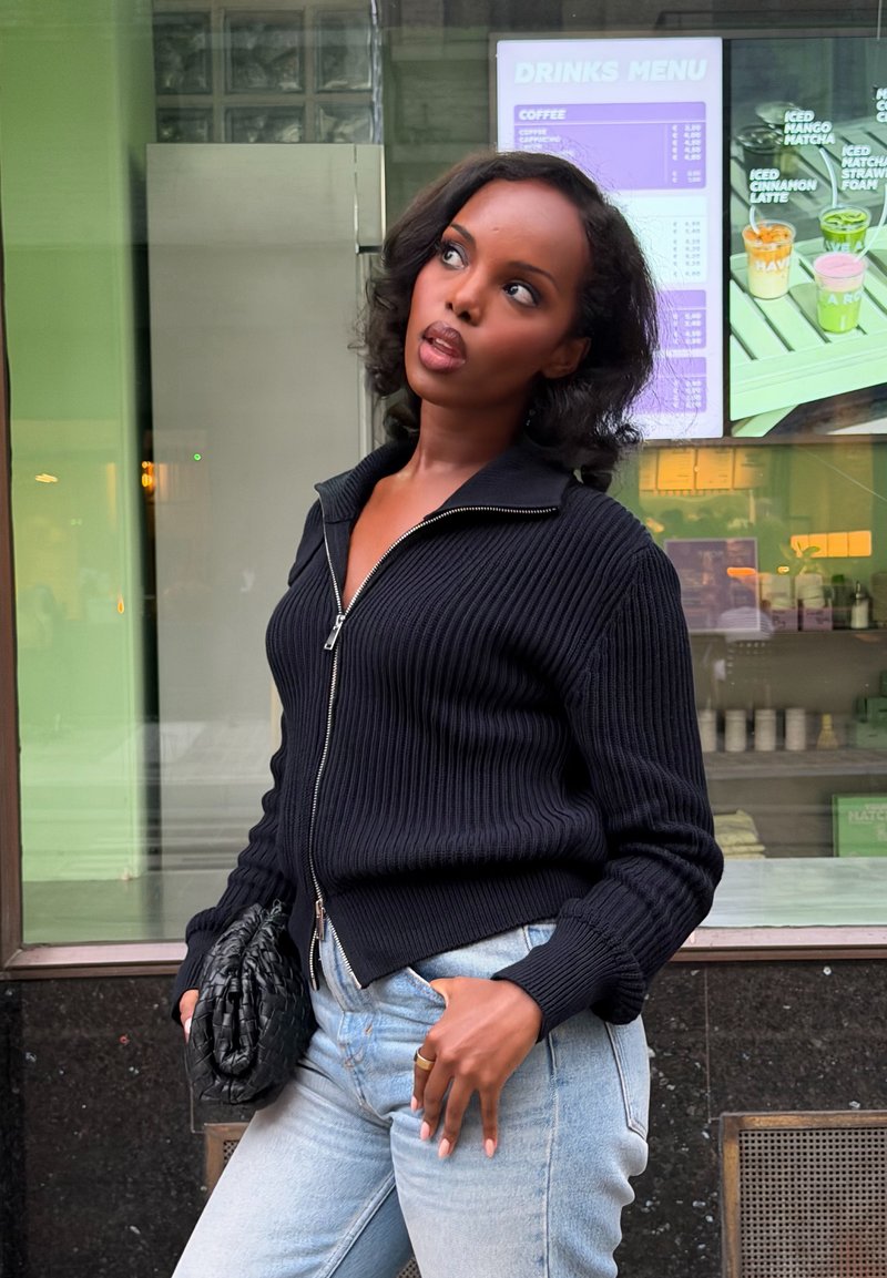 Mujer con un suéter negro con cremallera y vaqueros claros que sostiene un clutch negro, está cerca de una ventana con un menú de bebidas visible en el interior.