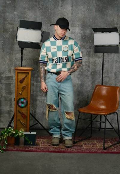 Hombre con camisa a cuadros, jeans rasgados y gorra negra, de pie entre dos luces de estudio, junto a una silla marrón y una vitrina de madera con objetos musicales.
