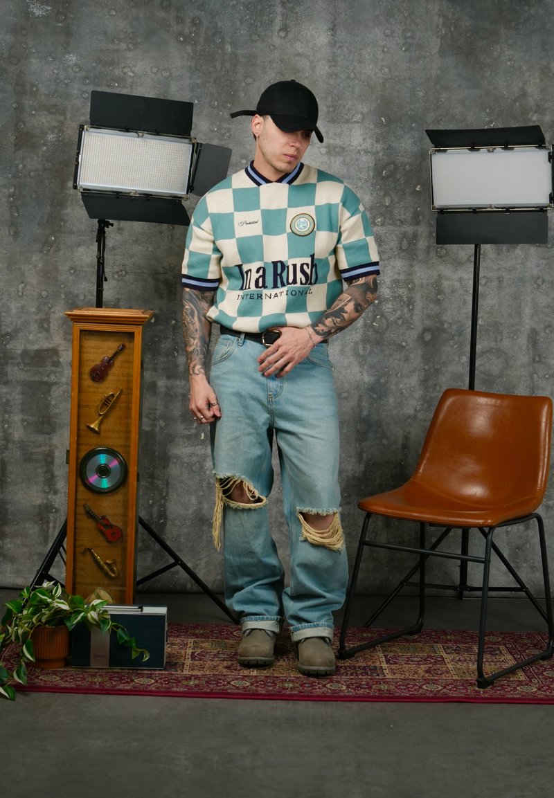 Bărbat îmbrăcat într-o cămașă în carouri, blugi rupți și șapcă neagră, stă între două lumini de studio, lângă un scaun maro și un suport din lemn cu obiecte muzicale.