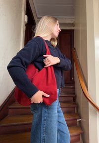 Γυναίκα με μακριά ξανθά μαλλιά που φοράει μαύρο πουλόβερ και τζιν κρατά μια μεγάλη κόκκινη τσάντα σε μια ξύλινη σκάλα μέσα σε εσωτερικό χώρο.