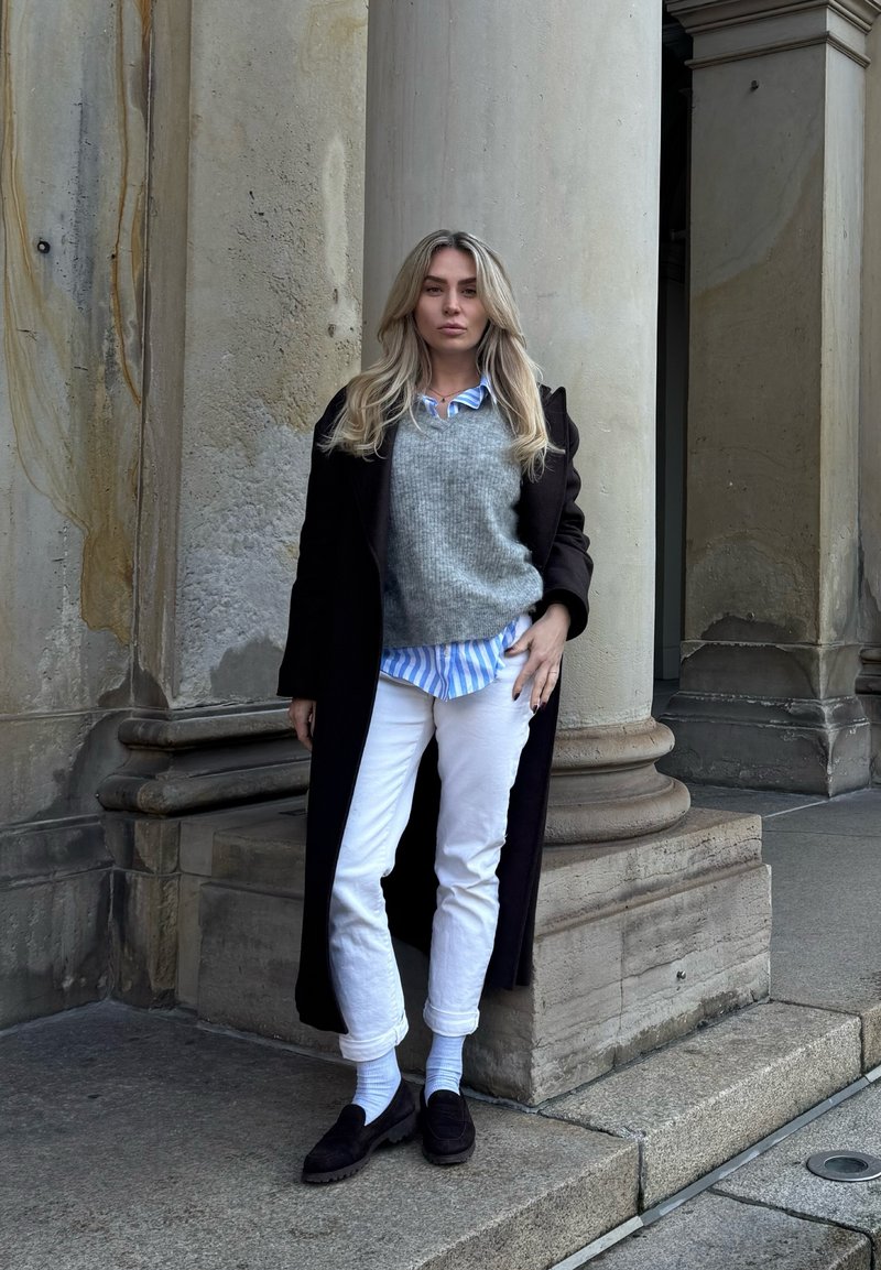 Long manteau noir, pull gris sur une chemise rayée bleue, pantalon blanc raccourci et mocassins foncés. Debout près de colonnes en pierre classiques.