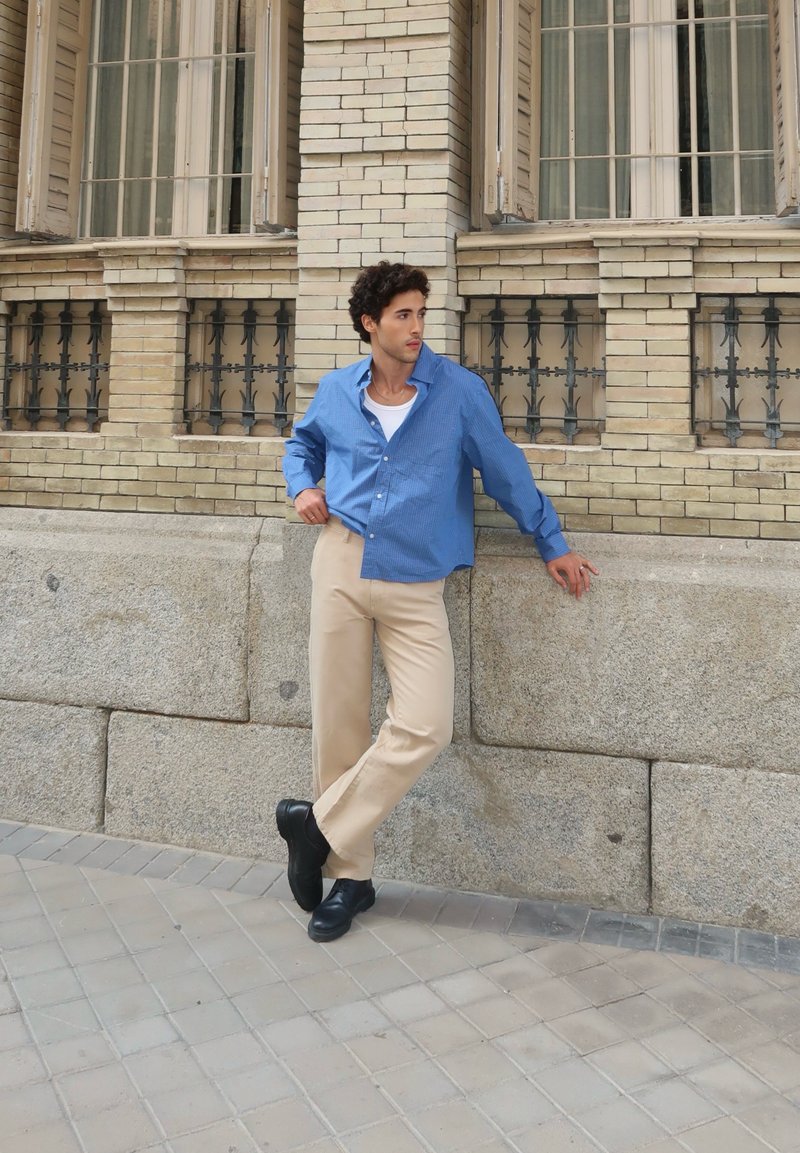 Camisa de botão xadrez azul, calças de creme e sapatos pretos. A camisa tem um corte descontraído e colarinho aberto. O fundo apresenta uma parede de pedra e janelas.