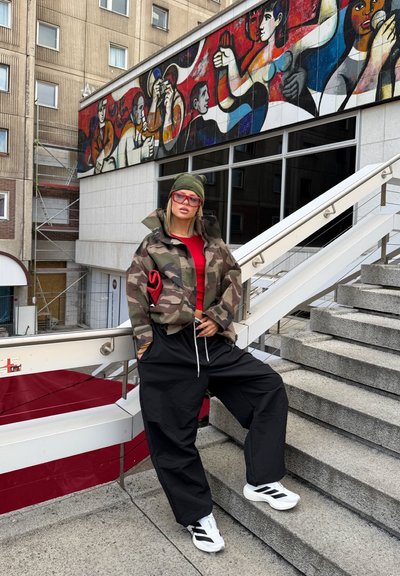 Chaqueta de camuflaje, parte superior roja, pantalones negros holgados y zapatillas blancas con rayas negras. Accesorio: gorro verde. Entorno urbano con un mural.