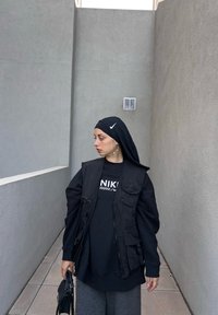 Μαύρο γιλέκο με φερμουάρ και πολλές τσέπες πάνω από μαύρη φόρμα Nike, συνδυασμένο με γκρι παντελόνι φάρδους και μαύρο χιτώνα. Περιλαμβάνεται μαύρη τσάντα.