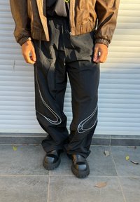Personne portant un pantalon large noir avec des motifs blancs incurvés, des bottes noires épaisses et une veste en cuir marron, debout sur un sol carrelé.