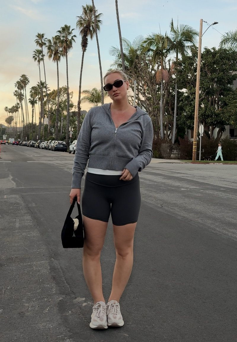 Frau mit Sonnenbrille, grauem Kapuzen-Sweatshirt mit Reißverschluss, schwarzen Shorts, weißen Turnschuhen, hält eine schwarze Tasche, steht an einer von Palmen gesäumten Straße bei Sonnenuntergang.