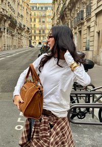 Smeđa nubuk torba s dvostrukim ručkama, s zlatnim akcentima i detaljima na zipu. U kombinaciji s bijelom košuljom dugih rukava i kariranim suknjom.