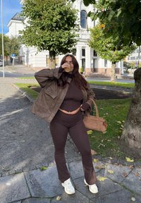 Brązowa skórzana kurtka na brązowym krótkim topie i dzwonowatych spodniach, trzymająca kubek, z brązową torbą i białymi trampkami. Tło z zielenią i budynkami.